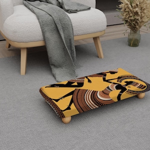 May include: A rectangular footstool with a yellow background and a black and brown tribal design featuring two figures. The footstool has rounded corners and four wooden legs. The footstool is placed on a grey carpet.