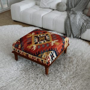Peut inclure: Un pouf carré doté d'un dessus en textile tissé aux couleurs vives rouge, bleu et jaune avec un motif géométrique. Le meuble est soutenu par quatre pieds courts en bois brun foncé, offrant une base stable à cet élément de mobilier bas.