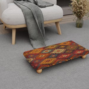 May include: A rectangular footstool with a vibrant geometric pattern in red, orange, yellow, and blue. It has four short, light-coloured wooden legs. The design is similar to a traditional rug, and is suitable for a living room.