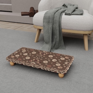 May include: A rectangular footstool with a brown floral pattern, resting on four small wooden legs. The footstool is placed on a gray rug, with a white armchair and a gray blanket in the background.