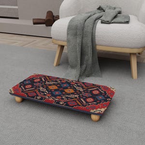 May include: A rectangular footstool with a patterned design in red, blue, and gold. The footstool has a dark blue border and is supported by four small, rounded wooden legs. The design features geometric shapes and floral motifs.
