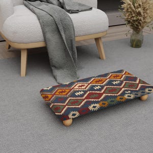 May include: A rectangular footstool with a colourful geometric pattern in shades of blue, red, orange, and white. The footstool has four short, light-coloured wooden legs. The footstool is placed on a grey rug.