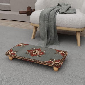 May include: A rectangular footstool with a gray background and a floral pattern in red and cream. The footstool has four rounded wooden legs. A gray blanket and chair are in the background.