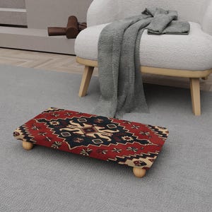 May include: A rectangular footstool with a red, black, and beige patterned design. The footstool has four rounded wooden legs. The design features geometric shapes and floral motifs. The footstool is placed on a grey carpet.