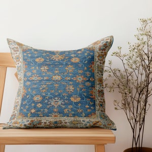 May include: A square cushion with a blue and orange floral design, placed on a wooden chair. The cushion features a detailed, vintage-inspired pattern with a border. The chair is made of light-coloured wood, and a plant with white flowers is next to it.