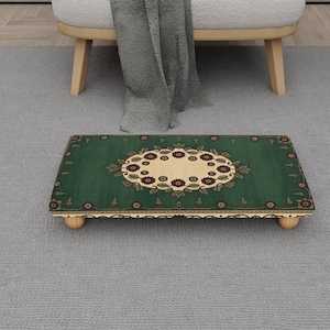 Puede incluir: Un reposapiés rectangular con una parte superior verde con un diseño floral en crema y marrón. El reposapiés tiene un borde decorativo y descansa sobre cuatro patas de madera pequeñas y redondeadas. El reposapiés está colocado sobre una alfombra gris.