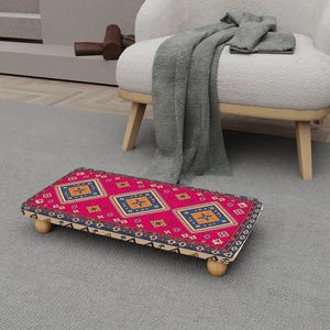 May include: A rectangular footstool with a vibrant pink and patterned top. The design includes blue, yellow, and white geometric shapes. The footstool is supported by four small, rounded wooden legs, and is placed on a gray carpet.