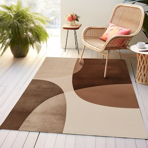 May include: A rectangular area rug featuring a large-scale abstract pattern with overlapping curved shapes in shades of dark brown, medium brown, and cream. The rug has a flat-weave texture and covers a portion of a white wooden floor surface.