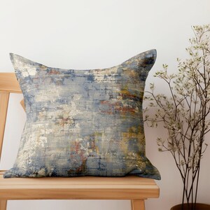 Puede incluir: Una almohada decorativa con un patrón abstracto en tonos azules, grises y marrones. La almohada está sentada en una silla de madera.