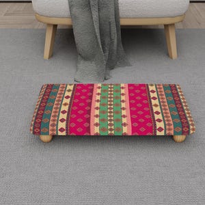 May include: A rectangular footstool with a vibrant, patterned design. The top features a repeating geometric pattern in shades of pink, green, and beige. The footstool rests on four small, rounded wooden legs.