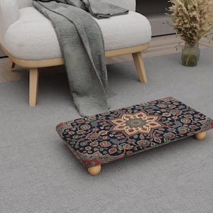 Könnte beinhalten: Ein rechteckiger Fußhocker mit einem dunkelblauen Muster mit einem zentralen Sterndesign in Rot, Gold und Grün. Der Hocker hat vier kurze, hellfarbige Holzbeine und steht auf einem grauen Teppich.