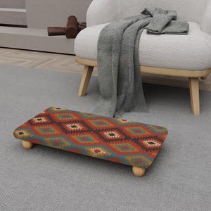 Puede incluir: Un reposapiés rectangular con un colorido patrón geométrico en rojo, naranja, verde azulado y marrón. El reposapiés tiene cuatro patas cortas de madera redondeadas. El diseño presenta formas de diamante y está colocado sobre una alfombra gris.