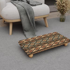 May include: A rectangular footstool with a geometric pattern in shades of brown, red, and green. The footstool has four small, rounded wooden legs. The design is reminiscent of a traditional rug.
