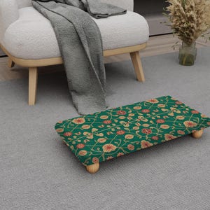 May include: A rectangular footstool with a green floral pattern featuring red and gold flowers and vines. The footstool has four short, light-coloured wooden legs. The footstool is placed on a grey carpet near a white armchair.