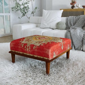Peut inclure: Un pouf carré doté d'un revêtement en tissu rouge vif orné d'un motif médaillon détaillé beige et doré. Le meuble repose sur quatre pieds en bois fuselés d'une finition brun moyen, offrant une base stable pour l'assise rembourrée.