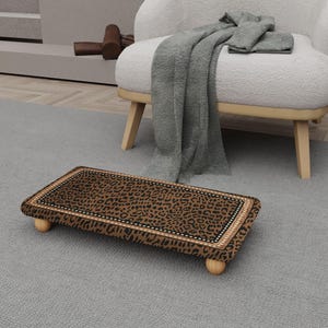May include: A rectangular footstool with a leopard print design. The stool has a brown and black pattern, a decorative border, and four small, rounded wooden legs. The footstool is placed on a gray carpet.