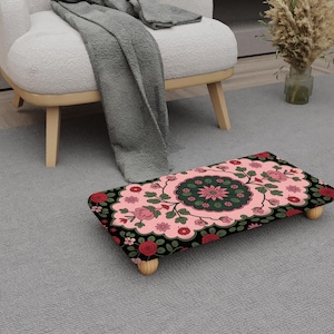 May include: A rectangular footstool with a floral pattern. The top has a pink base with a central green and pink floral medallion, framed by red roses and green leaves. It has four wooden legs.