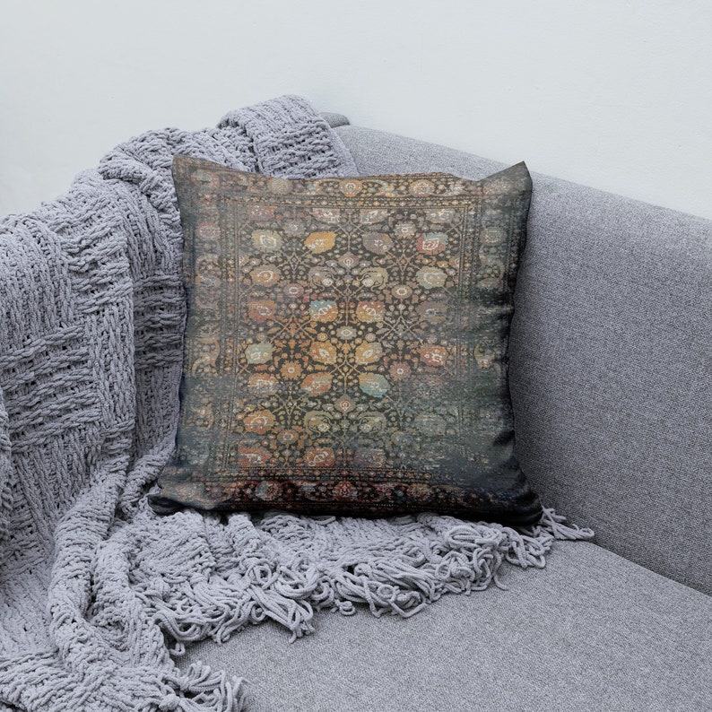 K&ouml;nnte beinhalten: Ein dekoratives quadratisches Kissen mit einem Vintage-Teppichdesign. Das Kissen hat einen dunklen Hintergrund mit einem sich wiederholenden Blumenmuster in Orange-, Gelb- und Blaut&ouml;nen. Es liegt auf einem grauen Sofa mit einem grauen Strickwurf.
