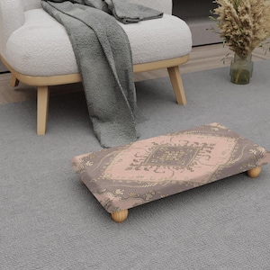 May include: A rectangular footstool with a pink and brown patterned fabric top and four small wooden legs. The footstool is placed on a grey carpet, near a white armchair and a grey blanket.