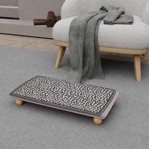 May include: A rectangular footstool with a leopard print top and light wood legs. The edges are trimmed with a black and white border. The footstool is on a gray rug.