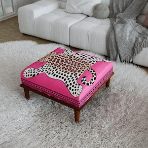 Puede incluir: Un otomano cuadrado con una cubierta de tela rosa vibrante que presenta un estampado de leopardo estilizado en blanco y negro. La pieza se apoya sobre cuatro patas cortas de madera marrón oscuro, proporcionando una base estable y baja.