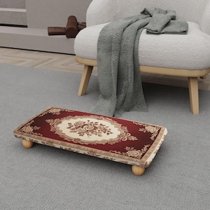 Op de afbeelding: Een rechthoekige voetenbank met een bordeauxrood en crèmekleurig bloemmotief. De voetenbank heeft een crèmekleurige rand en wordt ondersteund door vier kleine, afgeronde houten poten. Het bloemmotief is gecentreerd op het bovenoppervlak.