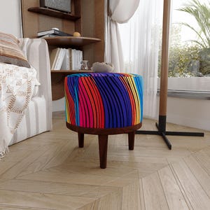 Pode incluir: Um banco redondo com um estofamento de tecido vibrante e multicolorido, apresentando um padrão de linhas verticais onduladas em tons de vermelho, azul, amarelo e preto. O assento é apoiado por quatro pernas de madeira castanho-escura.