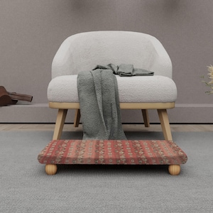 Può includere: Una scena d'interni accogliente con una poltrona bianca, una coperta grigia e un poggiapiedi a fantasia. Il poggiapiedi ha un design rosso e beige. La poltrona ha gambe in legno. Le piante secche in un vaso di vetro aggiungono un tocco naturale.
