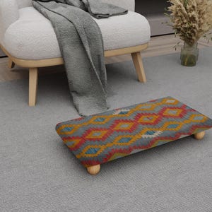 May include: A rectangular footstool with a gray background and a colorful geometric pattern in red, yellow, and blue. The footstool has four wooden legs and is placed on a gray rug.