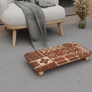 May include: A rectangular footstool with a patterned design in shades of brown, beige, and green. The footstool has four small, rounded wooden legs. The design features geometric shapes and patterns, creating a decorative accent piece.
