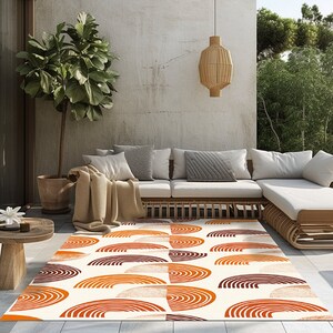 May include: A white area rug with a geometric pattern of orange and brown semi-circles. The rug is laid out on a patio with a white couch and a potted plant.