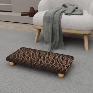 May include: A rectangular footstool with a brown and tan leopard print pattern. The footstool has rounded wooden legs. The footstool is on a gray rug, near a white armchair with wooden legs.