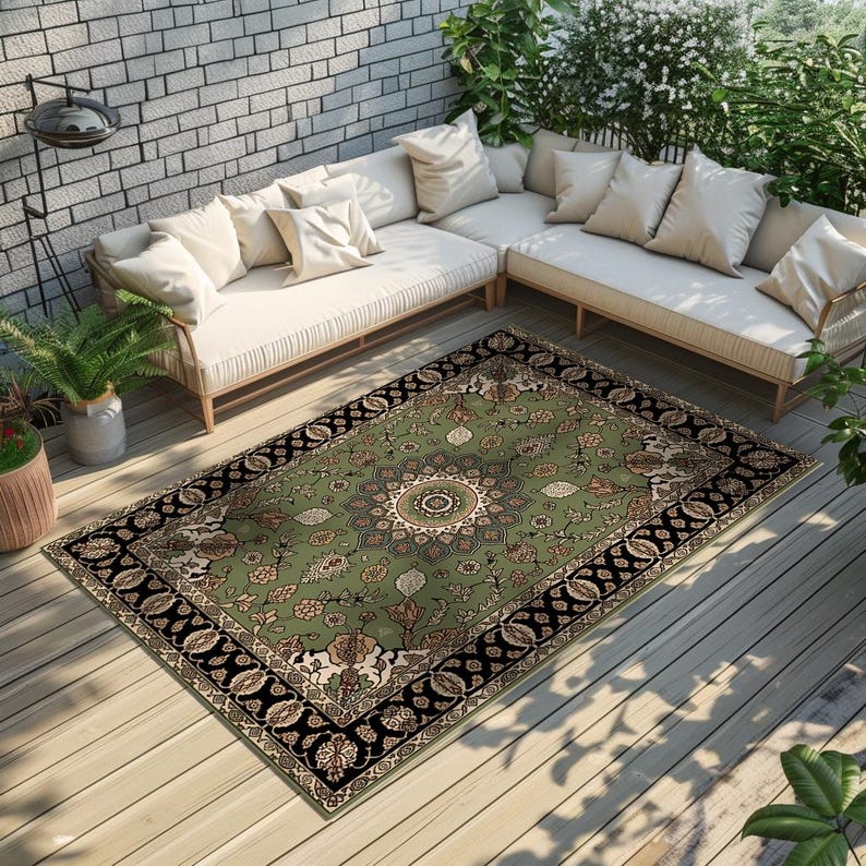 a cozy outdoor seating area with a large, ornate rug on a wooden deck. The seating area consists of a white sofa and a love seat, both adorned with white cushions. The area is surrounded by lush greenery, including several potted plants, creating a serene and inviting atmosphere.