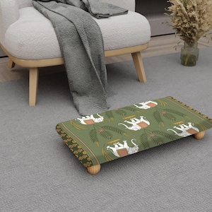 May include: A rectangular footstool with a green background and a pattern of white elephants and palm leaves. The footstool has rounded wooden legs and a decorative border. The stool is placed on a grey carpet.