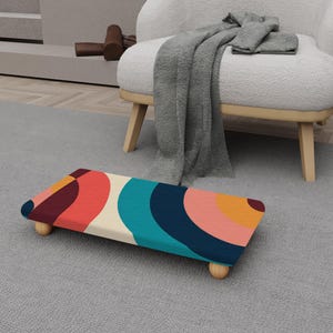May include: A rectangular footstool with a colorful abstract design. The top features a pattern of overlapping curved shapes in shades of red, orange, teal, and navy blue. The stool rests on four small, rounded wooden legs.