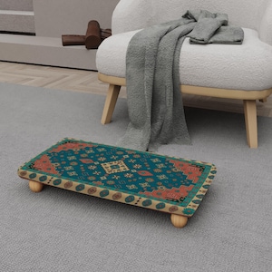 Puede incluir: Un reposapiés rectangular de madera con una parte superior estampada en turquesa y naranja. El diseño presenta formas geométricas y motivos florales. El taburete tiene cuatro patas de madera redondeadas y está sobre una alfombra gris.