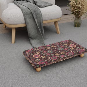 May include: A rectangular footstool with a dark brown floral pattern. The pattern includes pink, teal, and yellow flowers and leaves. The footstool has four short, light-colored wooden legs. The footstool is placed on a gray carpet.