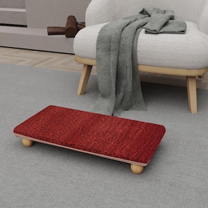 May include: A rectangular footstool with a red leopard print fabric top. The footstool has a cream-colored trim and is supported by four small, rounded wooden legs. The footstool is placed on a gray carpet.
