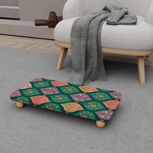 Puede incluir: Un reposapiés rectangular con un colorido estampado floral en tonos verdes, naranjas, rosas y morados. El reposapiés tiene cuatro patas cortas de madera redondeadas y está colocado sobre una alfombra gris.