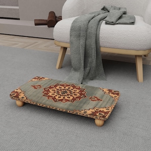 May include: A rectangular footstool with a patterned design in shades of teal, brown, and gold. The footstool has rounded wooden legs and is placed on a gray rug. The design features a central medallion and border details.