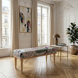 Puede incluir: Un banco decorativo con un diseño floral y de pavo real. El banco tiene patas de madera clara y forma rectangular. La habitación también cuenta con un cuadro grande, una mesa auxiliar con una lámpara y una planta en maceta.