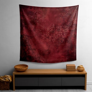 May include: A red tapestry with a distressed, vintage look hangs on a white wall above a wooden shelf with a bowl and books.