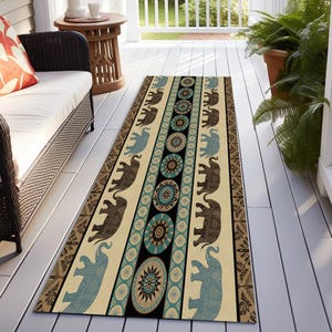 May include: A rectangular outdoor rug with an elephant pattern. The rug features alternating rows of brown and blue elephants on a cream background, with a central stripe of teal and black circular designs. The border has a brown floral pattern.