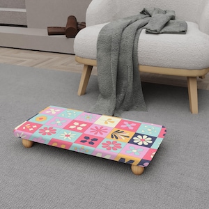 Puede incluir: Un reposapiés rectangular con un colorido estampado floral en cuadrados rosas, azules, amarillos y azul marino. El reposapiés tiene patas de madera redondeadas y está colocado sobre una alfombra gris. El diseño presenta varios motivos de flores y hojas.