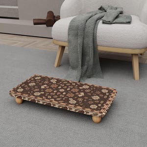 May include: A rectangular footstool with a dark brown floral pattern. The footstool has a light brown border and is supported by four small, rounded wooden legs. The design features a repeating pattern of flowers and leaves.
