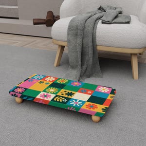 May include: A colorful footstool with a floral pattern in a patchwork design. The footstool has a rectangular shape and is supported by four small, rounded wooden legs. The pattern includes flowers in various colors, including pink, yellow, and green.