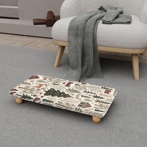 May include: A rectangular footstool with a nature-themed pattern featuring trees, houses, and animals in shades of brown, green, and red on a cream background. The footstool is supported by four small wooden legs.