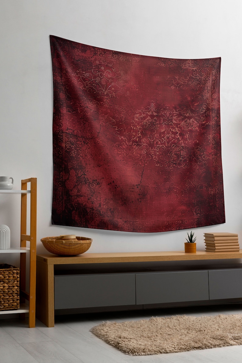 May include: A red tapestry with a faded floral pattern hangs on a white wall above a wooden shelf and a dark gray media console with three drawers. A small potted plant and a stack of books are on the console.