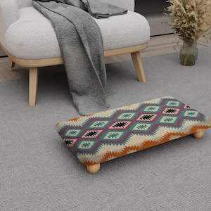 Peut inclure: Un repose-pieds rectangulaire avec un motif géométrique coloré en turquoise, violet, orange et noir sur fond crème. Le repose-pieds a quatre pieds courts en bois clair et est posé sur un tapis gris.