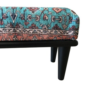 A colorful, patterned ottoman or footstool with a teal and orange color scheme, placed on a black wooden frame.